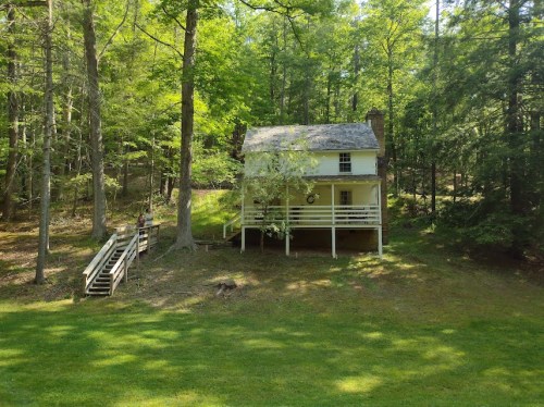 A small yellow house surrounded by trees, with a wooden staircase leading up to it and a grassy area in front.