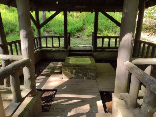 A wooden gazebo with a stone platform in the center, surrounded by greenery and wooden railings.