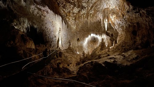 A dimly lit cave with stalactites and stalagmites, featuring a winding path and a person exploring the rocky terrain.