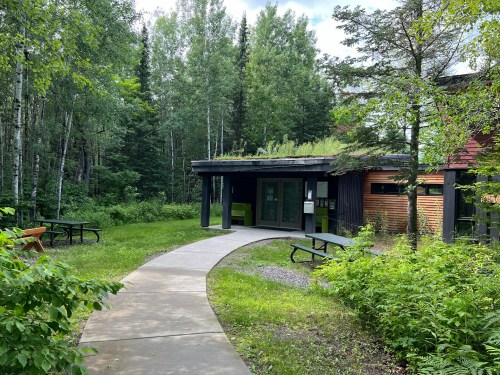 A winding path leads to a modern building surrounded by lush greenery and trees. Picnic tables are visible nearby.