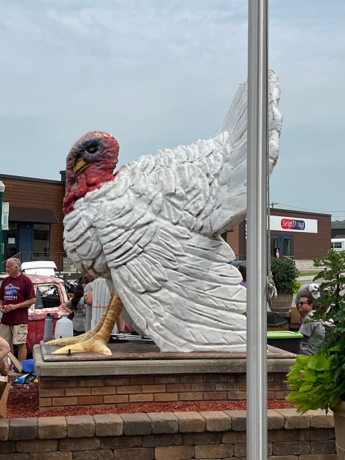 A large, white turkey statue stands in a plaza, surrounded by people and greenery on a cloudy day.