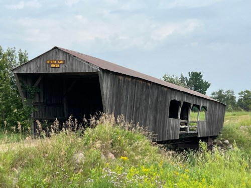A weathered wooden covered bridge overgrown with grass, labeled "Otter Tail River," under a cloudy sky.