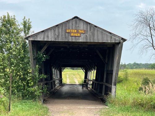 A wooden covered bridge with a sign reading "Otter Tail River," leading to a grassy area and cloudy sky.