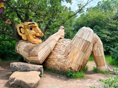 A large wooden sculpture of a reclining figure surrounded by greenery, made from various wooden planks.