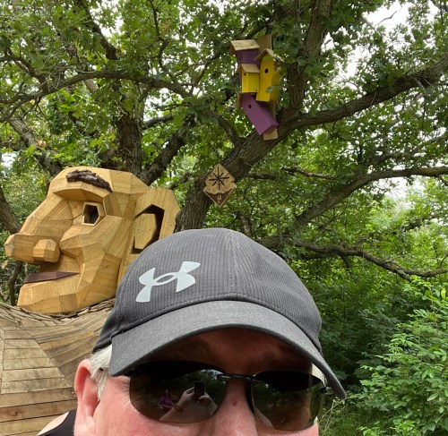 A person wearing a cap and sunglasses takes a selfie in front of a large wooden sculpture of a head in a wooded area.