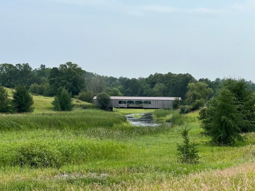 A covered bridge spans a calm stream, surrounded by lush green fields and trees under a cloudy sky.