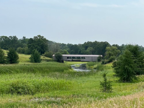 A covered bridge spans a calm river, surrounded by lush green fields and trees under a cloudy sky.