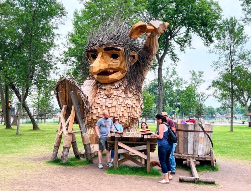 A large wooden troll sculpture stands in a park, with people interacting around it and trees in the background.