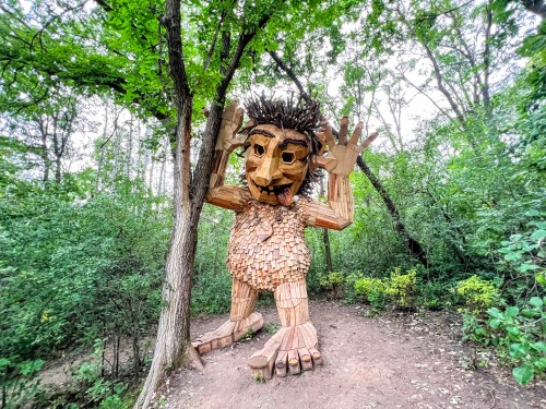 A large wooden sculpture of a troll with a playful expression, surrounded by trees and greenery in a forest setting.