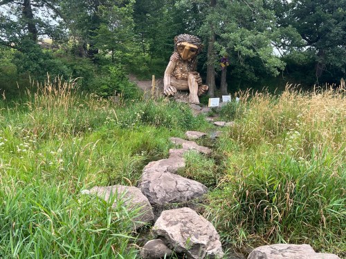 A large wooden troll sculpture sits among tall grass and trees, with a stone path leading towards it.