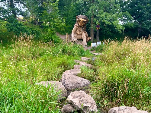 A large wooden troll sculpture sits among tall grass and trees, with a stone path leading towards it.