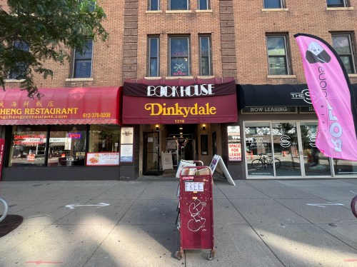 A storefront with a red awning reading "Dinkydale" and "Bookhouse," surrounded by other businesses and a free book cart.