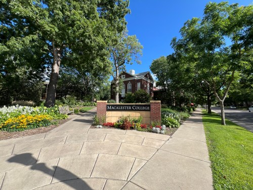 Sign for Macalester College surrounded by greenery and flowers on a sunny day. Pathway leads to the college entrance.