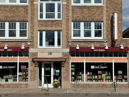A brick building with large windows, featuring a bookstore named "next chapter" with colorful displays and an open sign.