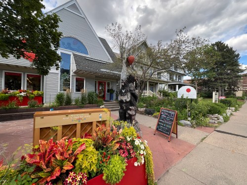 A charming street scene featuring a wooden sculpture, colorful flowers, and a sign outside a quaint building.