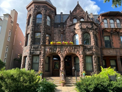 A historic stone building with ornate architecture, large windows, and flower boxes, surrounded by greenery.