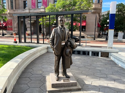 A bronze statue of a man in a suit holding a hat, standing on a stone pedestal in a public plaza.