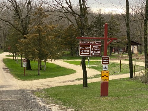 Signpost at a river access point with directions for boat ramp, kayak, and canoe entrances, surrounded by trees.