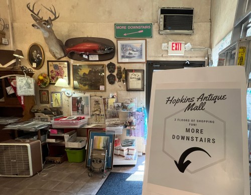 Interior of an antique shop with a sign directing to more items downstairs, featuring various vintage decor and artwork.