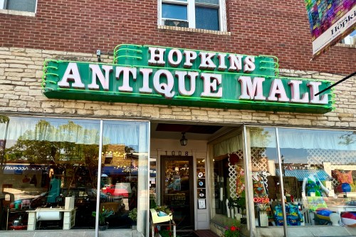 Sign for "Hopkins Antique Mall" with a green neon border, showcasing a vintage storefront with large windows.