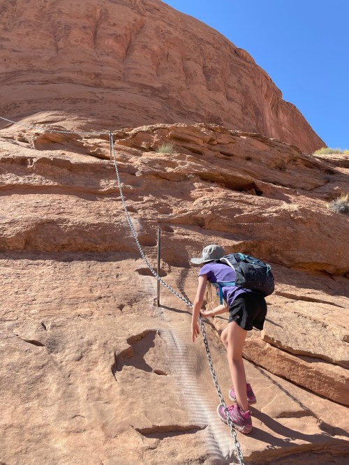 A child climbs a rocky trail with a chain for support, surrounded by red rock formations under a clear blue sky.