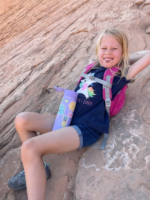 A smiling girl in a backpack relaxes on a rock, holding a colorful water bottle, enjoying the outdoors.