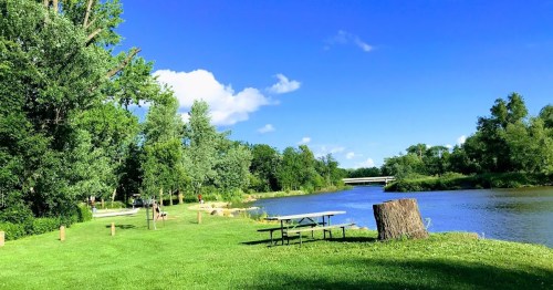 A serene riverside scene with green grass, trees, a picnic table, and a clear blue sky.
