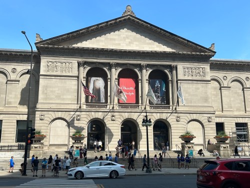 The Art Institute of Chicago's grand facade with banners, flags, and visitors outside on a sunny day.