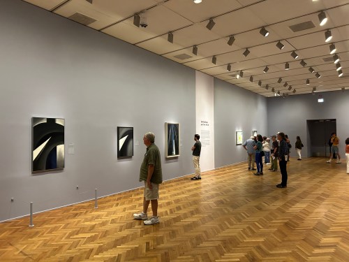 Visitors admire abstract paintings in a spacious art gallery with wooden flooring and soft lighting.