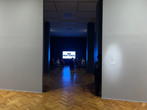 A gallery view showing a dark room with a screen displaying "HER NEW YORK" and people seated in front of it.