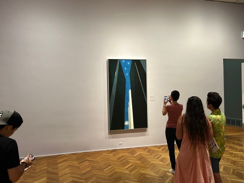 Visitors admire a tall painting of a narrow space with blue sky, surrounded by dark vertical shapes in an art gallery.