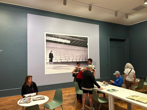 A gallery scene with visitors admiring a large black-and-white photo on the wall, surrounded by tables and chairs.