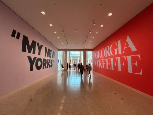 A spacious gallery with large text on walls: "MY NEW YORKS" on the left and "GEORGIA O'KEEFFE" on the right. Visitors walk through.