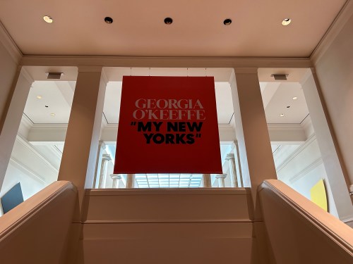 A large sign reading "Georgia O'Keeffe: My New Yorks" hangs above a staircase in a bright, modern gallery space.