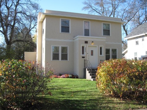 A two-story beige house with white trim, surrounded by green grass and colorful shrubs.