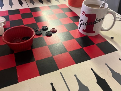 A checkers board with red and black pieces, a red bowl, and a "#1 Grandma" mug filled with coffee.