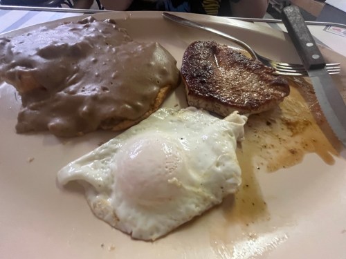 A plate with a fried egg, a piece of meat, and a biscuit topped with gravy. Fork and knife are beside the plate.