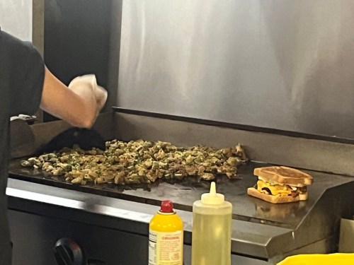 A cook prepares chopped ingredients on a grill, with a sandwich and condiments nearby.
