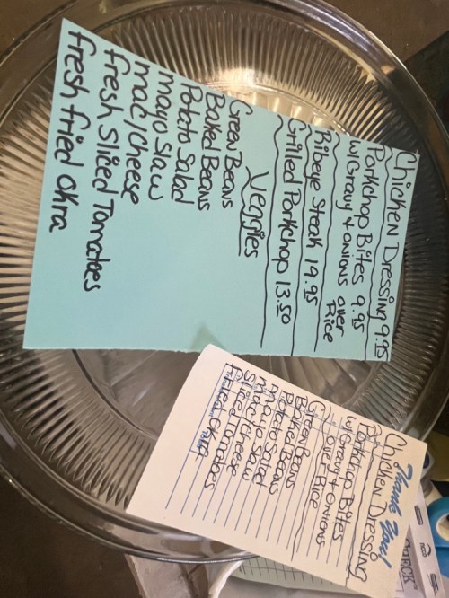 Menu items written on blue paper, listing chicken and dressing, pork chop, vegetables, and sides like fried okra and potato salad.
