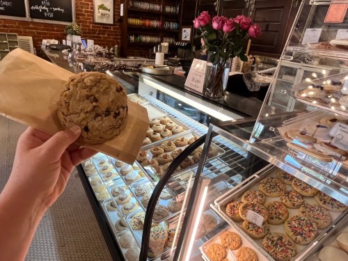 A hand holds a cookie in front of a display case filled with various cookies and a vase of roses.