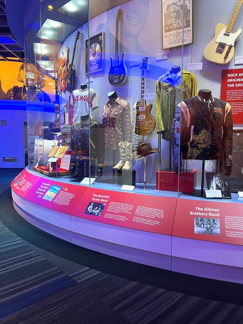 Display case featuring musical instruments, clothing, and memorabilia from the Allman Brothers Band and related artists.