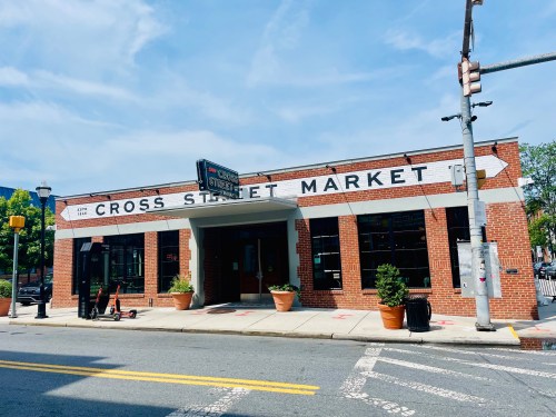 Exterior of Cross Street Market, a brick building with large windows and planters, under a clear blue sky.