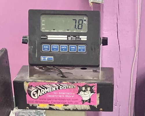 Digital scale displaying a weight of 7.8 kg, mounted on a stand with a colorful sticker for The Garment District.