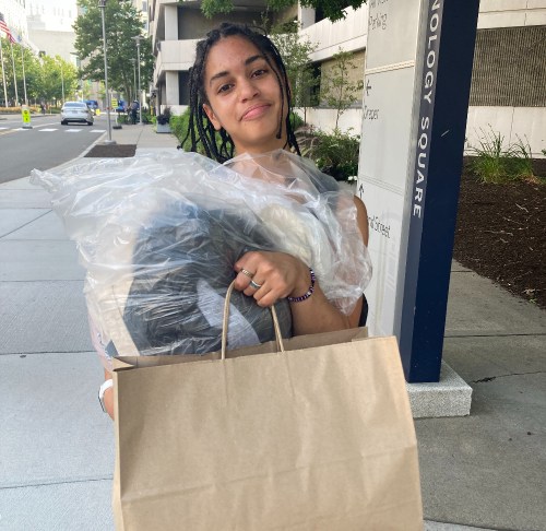 A young person with braided hair carries a large bag and a bundle wrapped in plastic on a city sidewalk.