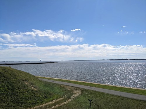 A serene view of a calm lake under a clear blue sky, with distant land and a path along the water's edge.