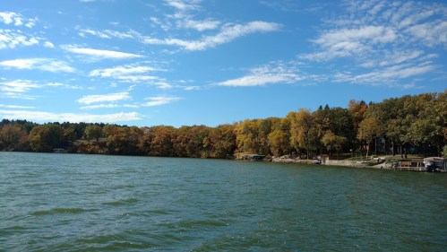 A serene lake scene with colorful autumn trees lining the shore under a blue sky with scattered clouds.