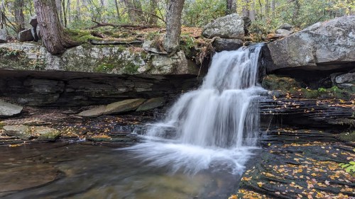 A serene waterfall cascades over rocks into a calm pool, surrounded by autumn foliage and lush greenery.