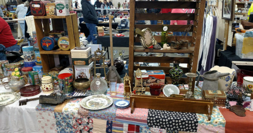 A table filled with various vintage items and antiques at a bustling market, with people browsing in the background.