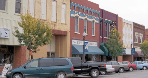A street view of a small town with colorful storefronts and parked cars along the curb. Trees line the sidewalk.