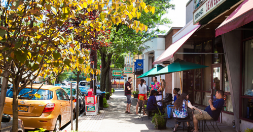 A vibrant street scene with people socializing, outdoor seating, and colorful storefronts under sunny skies.